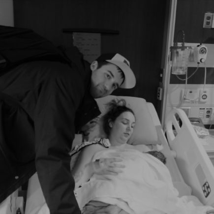 A man leans over his wife, who is is lying in a hospital bed with her their new baby.