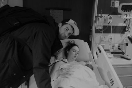 A man leans over his wife, who is is lying in a hospital bed with her their new baby.