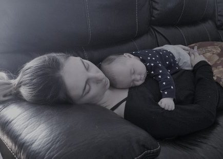 A mother sleeps on the couch with her baby, also sleeping, on her chest.