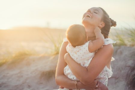 A woman holds her baby and smiles. The sun is shining in the background.
