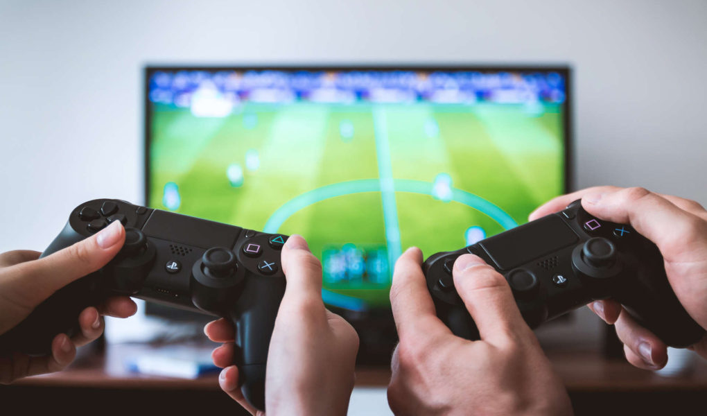 Two pairs of hands are holding PS4 controllers in front of a TV.