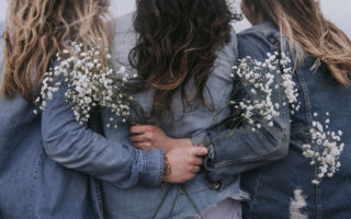 Three woman have their backs to the camera, and are embracing each other with flowers in their hands.