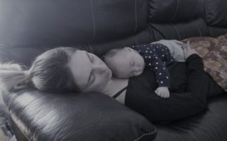 A mother sleeps on the couch with her baby, also sleeping, on her chest.
