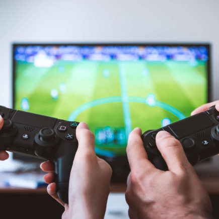 Two pairs of hands are holding PS4 controllers in front of a TV.