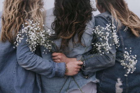 Three woman have their backs to the camera, and are embracing each other with flowers in their hands.