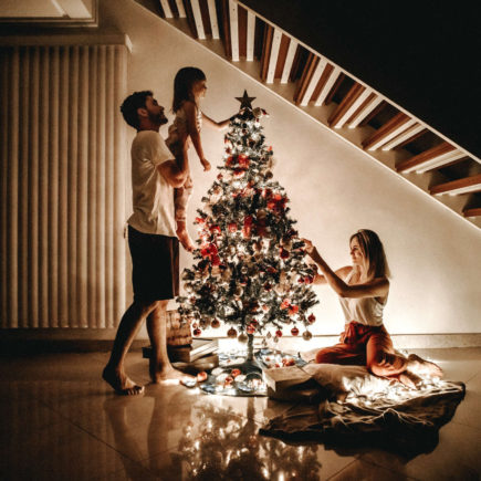 A small family is decorating a Christmas tree. Mom is on the ground and Dad is lifting the child.