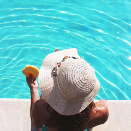A Woman is sitting on the edge of a pool, eating a piece of pineapple.